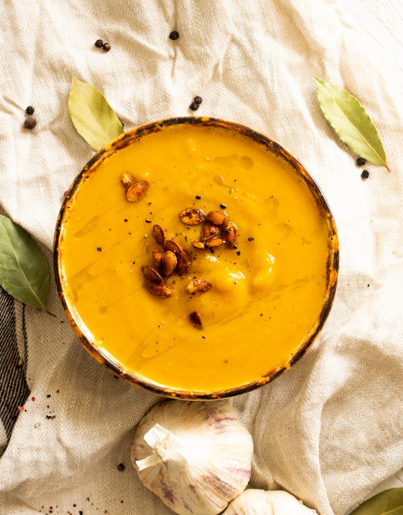 A picture of a creamy, vegan butternut squash soup from the top with a drizzle of olive oil and roasted pumpkin seeds on a linen cloth with garlic and spices